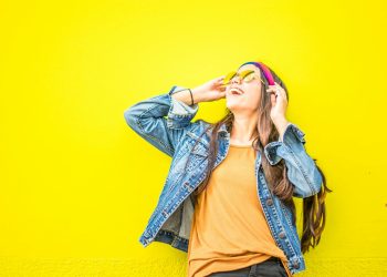 Smiling woman in sunglasses stands against vibrant yellow wall, radiating happiness.