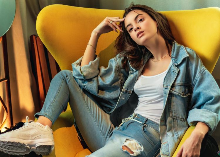 Stylish woman in denim poses on a modern yellow chair indoors.
