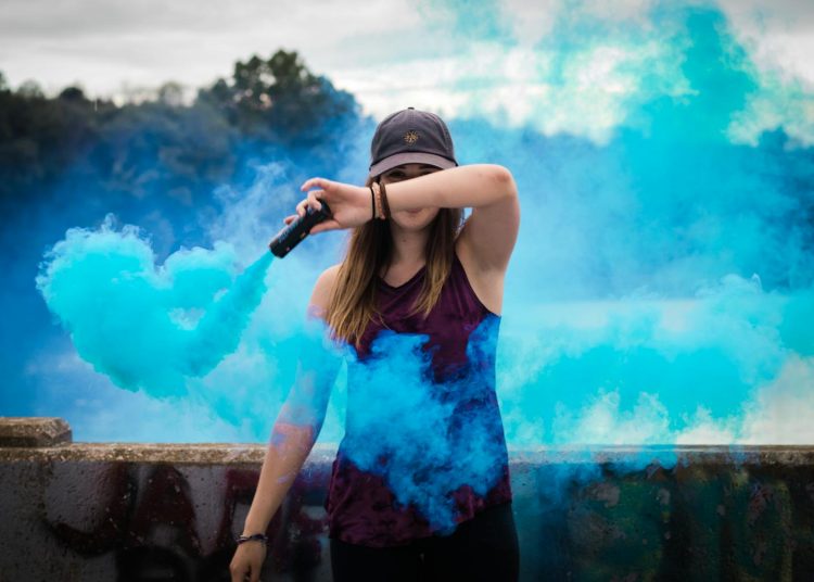 A stylish woman surrounded by bright blue smoke outdoors, creating a dynamic fashion statement.