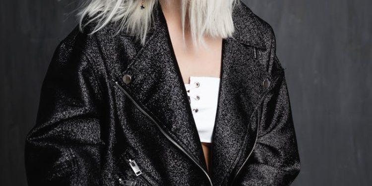 A fashionable young woman with blonde hair wearing a trendy jacket poses confidently indoors.