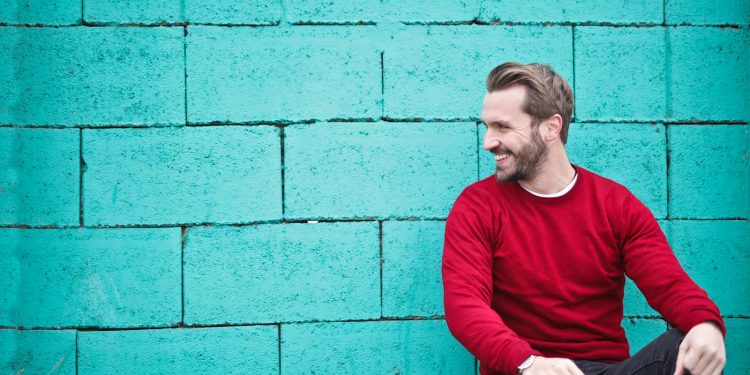 A cheerful man in a red sweater sits against a turquoise wall, enjoying a moment of happiness and laughter.