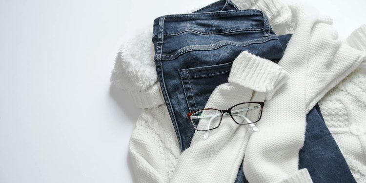 Stylish flat lay featuring denim jeans, white sweater, and eyeglasses on a bright background.